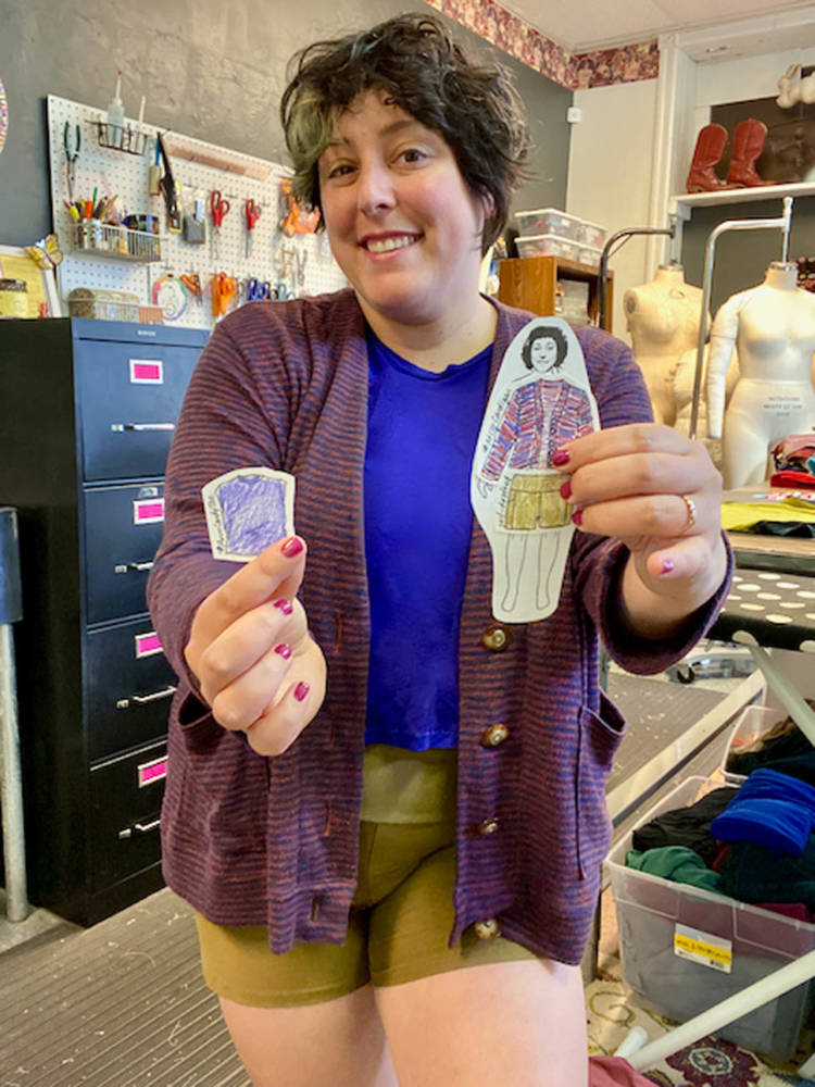 Indie pattern designer Ruby stands in her studio holding up a paper doll cutout of the My Body Model template dressed in a new addition to her handmade wardrobe. The paper doll wears a striped cardigan, olive green shorts, and a bright blue top. Ruby is wearing the same designs brought to life in fabric.