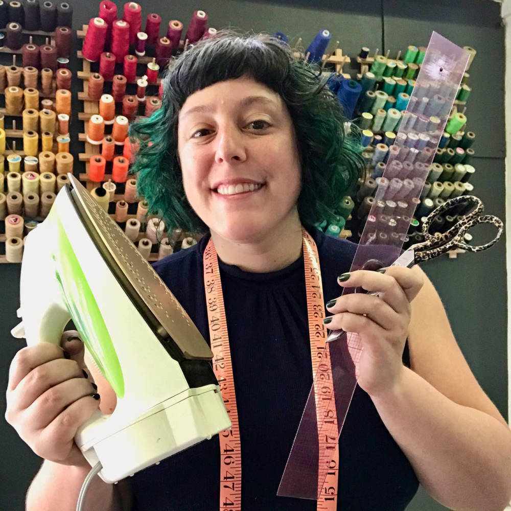 Ruby stands in front of a wall of rainbow thread, holding a pair of scissors, a clear plastic ruler, and an iron. She has a measuring tape draped around her shoulders, and her dark curly hair is dyed a vibrant teal-green.