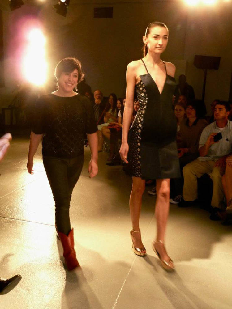 Two people are walking down a fashion runway together. The fashion model to the right is tall and slender and wears a silk cocktail dress. Ruby is on the left wears jeans, a sparkly black shirt, and red cowboy boots. Her body proportions are much shorter and wider than the model’s.