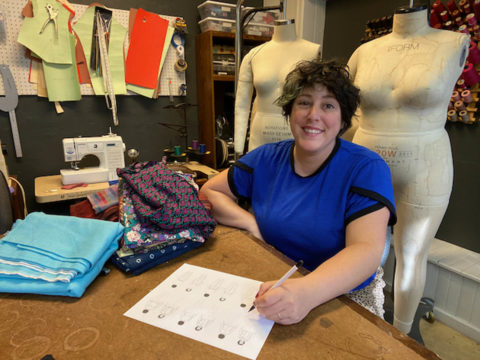 Fashion design with MyBodyModel custom croquis: Ruby sits at a table in her sewing studio surrounded by colorful thread, dress forms, and sewing machines. She is wearing a blue tee shirt made from her own Arm Candy Tee sewing pattern, and sketching on a sheet of paper that has six small My Body Model croquis figures on it.