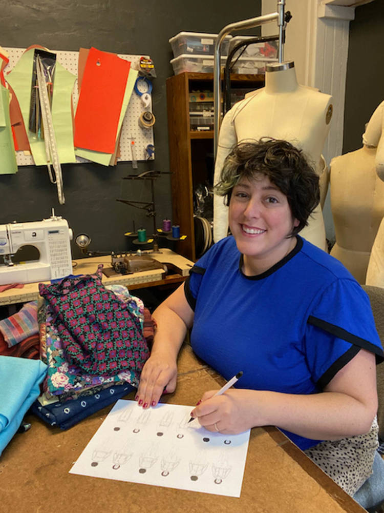 Fashion design with MyBodyModel custom croquis: Ruby sits at a table in her sewing studio surrounded by colorful thread, dress forms, and sewing machines. She is wearing a blue t-shirt made from her own Arm Candy Tee sewing pattern and sketching on a sheet of paper that has six small My Body Model figures on it.
