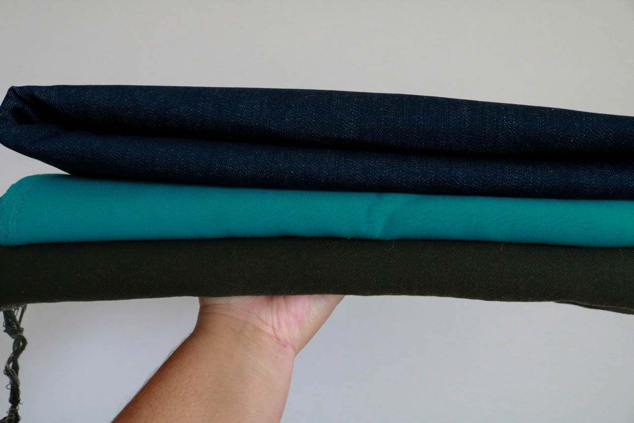 Amanda holds up a stack of fabrics from her sewing and knitting stash, including a dark indigo stretch denim, teal silk twill, and dark bull denim.