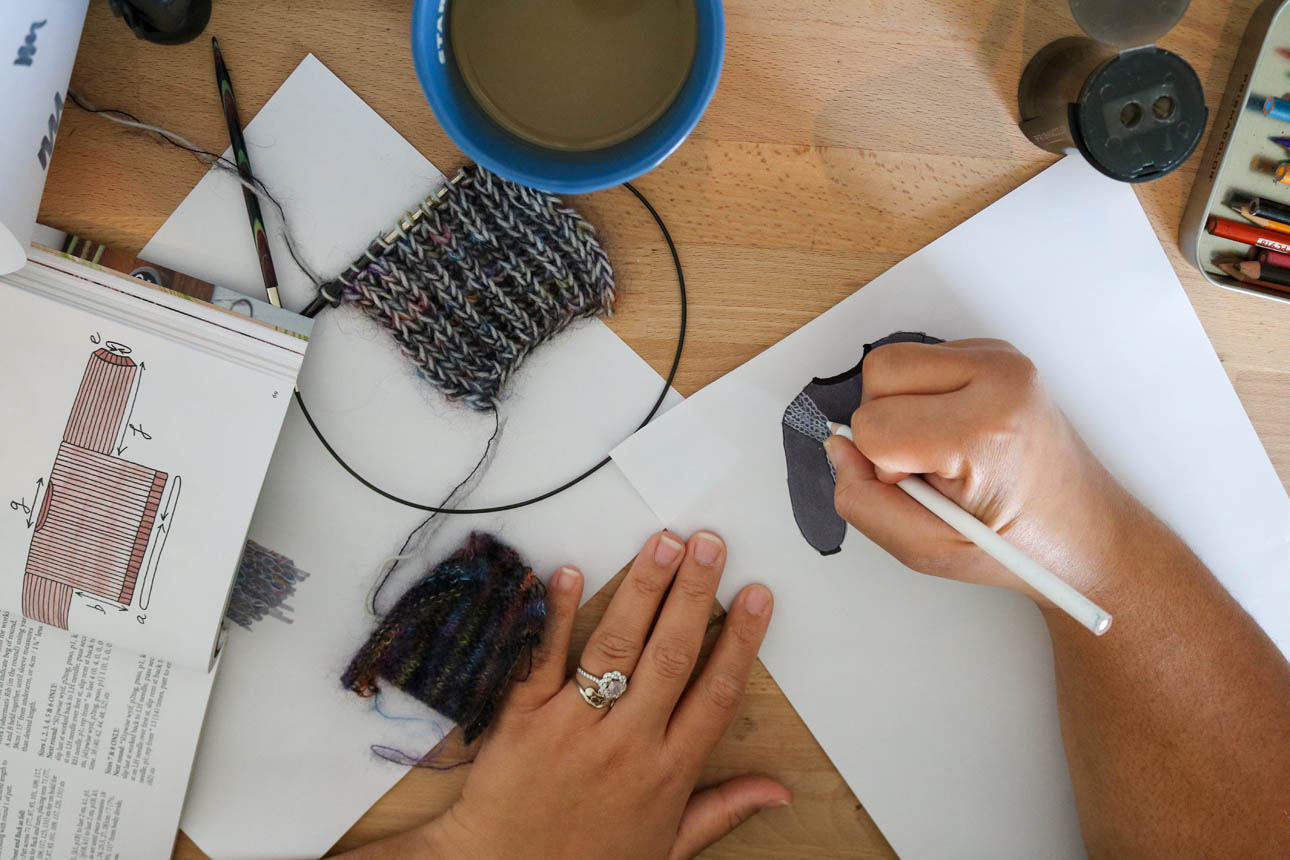 Amanda adds texture to a sketch of her Aloft Pullover sewing and knitting plans, drawn in gray using her MyBodyModel croquis, while surrounded by a coffee cup, knit fabric swatch, and art supplies.