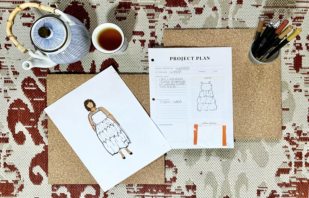 A spread of Tiffaney's MyBodyModel paper doll croquis wearing a paper garment version of the Amber dress from Seamwork alongside a teapot and cup of tea, cup of colored markers and pens, and a sheet for sewing project plans.