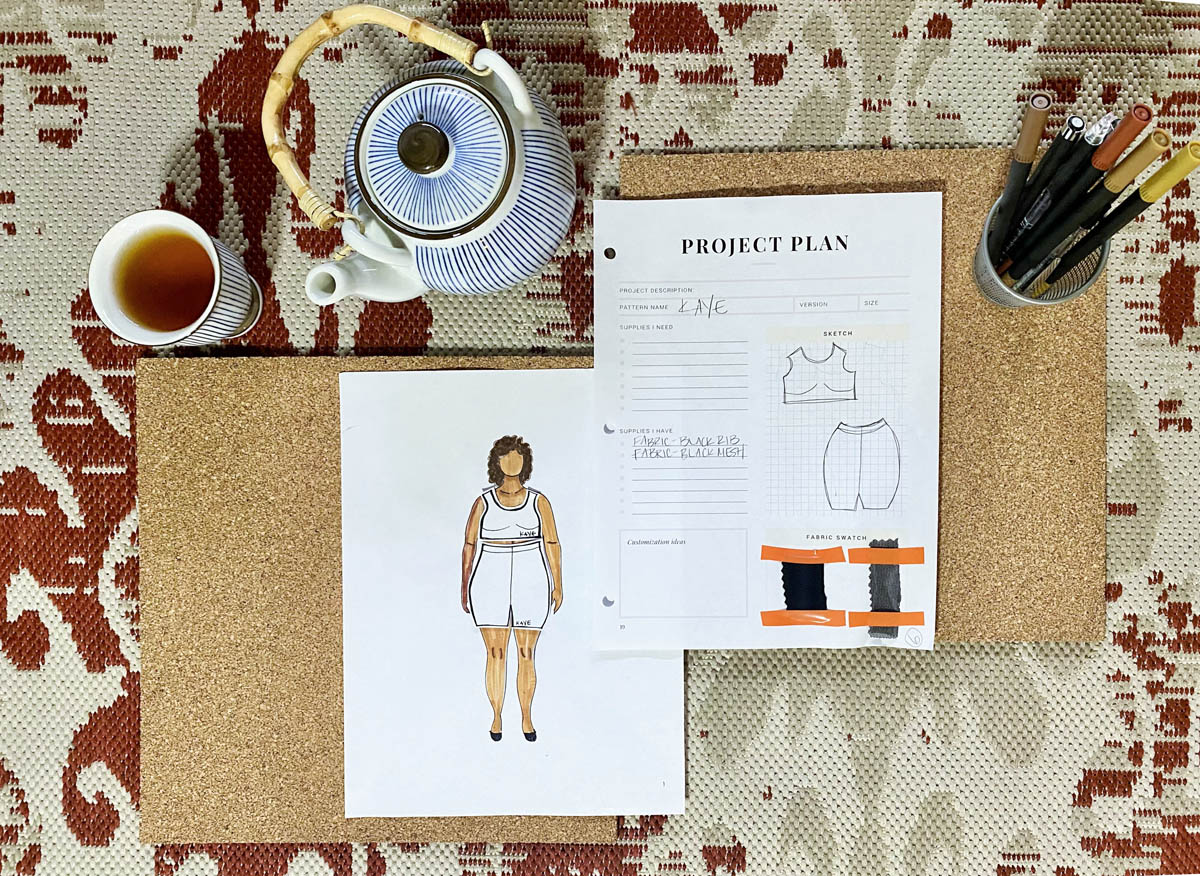 A spread of Tiffaney's MyBodyModel paper doll croquis wearing a paper garment version of the Kaye loungewear set from Seamwork alongside a teapot and cup of tea, cup of colored markers and pens, and a sheet for sewing project plans.